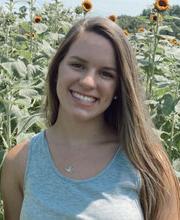 奥利维亚·迪恩，22岁, smiling with long straight light brown hair and wearing a light blue tank top with sunflowers in the background